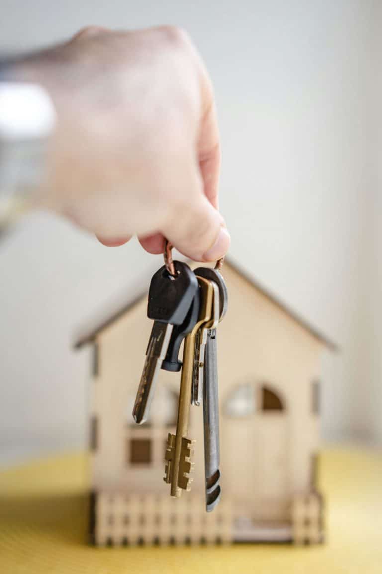 Une main tient un trousseau de clés devant une petite maquette de maison en bois. Les clés sont variées, avec des formes et des tailles différentes, indiquant qu’elles ouvrent différentes serrures. La maquette de la maison est posée sur une surface jaune. L’image évoque le concept d’achat ou de propriété d’une maison.